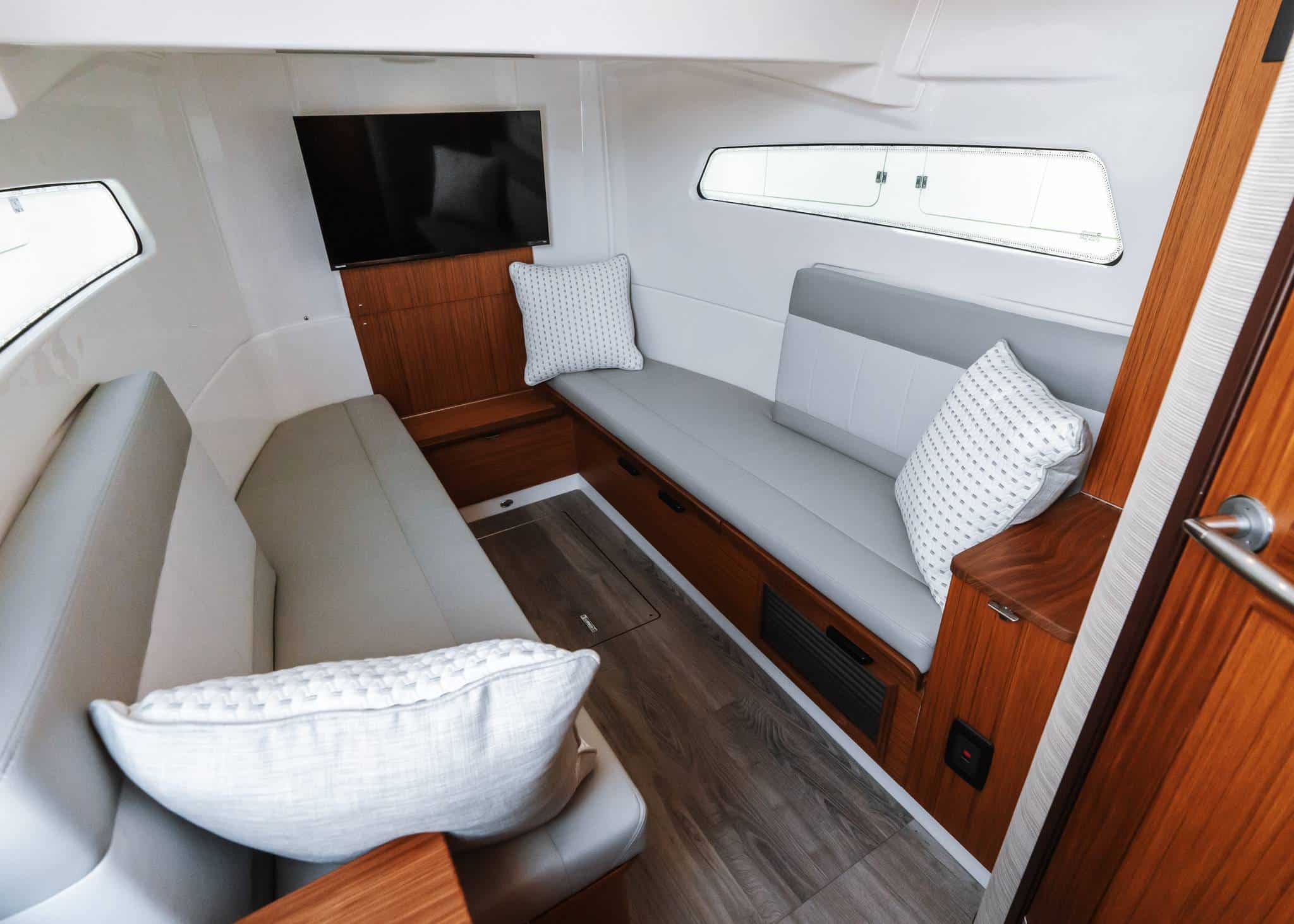 Interior of a modern Solace 415 CS luxury boat cabin featuring two cushioned benches, decorative pillows, wood accents, a wall-mounted TV, and windows that fill the space with natural light.